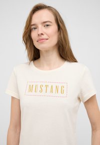Cream short-sleeve T-shirt made of soft cotton featuring a "MUSTANG" graphic in gold with decorative pink accents and a round neckline.