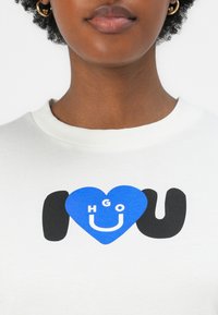 Witte sweatshirt met een ronde halslijn, versierd met een blauwe hartafbeelding die de tekst "HGO" bevat in vetgedrukte witte en zwarte letters "I" en "U" ernaast.