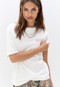 T-shirt en coton blanc à manches courtes, avec un logo brodé rose sur la poitrine gauche. Le mannequin porte des colliers en chaîne superposés et un pantalon à motifs.