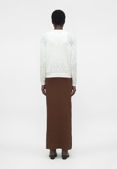 Άτομο που φοράει λευκό πουλόβερ με μακριά μανίκια και σχέδιο, καθώς και μακρύ καφέ φόρεμα, στέκεται γυρισμένο προς τα πίσω σε λευκό απλό φόντο.