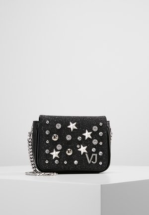 Sac bandoulière noir scintillant avec étoile argentée et décorations à clous ronds et une bandoulière en chaîne argentée sur une surface blanche.