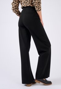 Pantalones anchos negros con una textura lisa y una cintura alta, con un bolsillo trasero. Combinados con mocasines negros gruesos.