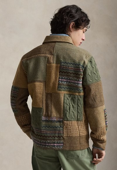 Άνδρας που φοράει πλεκτό πουλόβερ πατςγουορκ σε πράσινους και καφέ τόνους, όρθιος πλάγια με το κεφάλι γυρισμένο, με υφασματικά μοτίβα και γιακά ορατό.