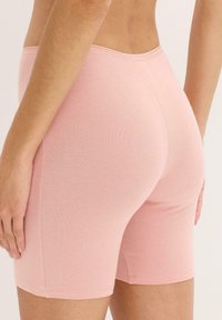 Gros plan d'une personne portant un short en tricot ajusté rose clair, montrant l'arrière et le côté de la taille jusqu'à mi-cuisse, avec une main détendue sur le côté.
