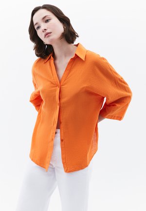 Orange struktureret skjorte med krave og drop-skuldere, kombineret med hvide bukser. Stoffet ser let og åndbart ud.