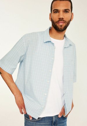 Mand med kort krøllet hår og skæg iført en lyseblå ternet kortærmet skjorte over en hvid t-shirt og blå jeans, hænderne i lommerne.