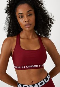 Mujer con cabello rizado vestida con un sujetador deportivo burdeos de Under Armour y leggings a juego, de pie frente a un fondo blanco liso.