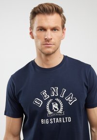 Tamsiai mėlynas medvilninis marškinėliai su priekinė spausdinimu, kuriame parašyta "DENIM" ryškiai baltais raidėmis, apsuptas laurų dizaino ir tekstu "BIG STAR LTD".