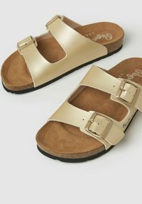 Sandalias beige sin talón con dos correas ajustables con hebillas, plantilla acolchada marrón, media suela de corcho y suela exterior negra sobre fondo blanco.