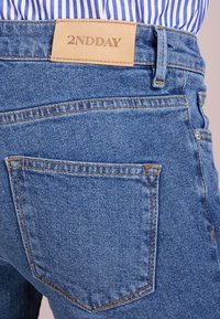 Vaqueros de mezclilla azul con un parche de cuero de la marca etiquetado "2NDDAY". Presenta un bolsillo trasero con costuras en contraste y un acabado texturizado.