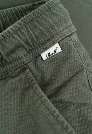 Pantalones cortos verdes de tela con cinturilla elástica, superficie texturizada y una pequeña etiqueta blanca con "Reell" cosida en ella.