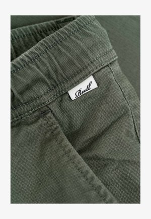 Pantalones cortos verdes de tela con cinturilla elástica, superficie texturizada y una pequeña etiqueta blanca con "Reell" cosida en ella.
