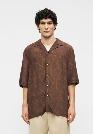 Άνδρας με κοντά σγουρά μαλλιά, που φοράει καφέ πουκάμισο με κοντά μανίκια και αφηρημένα σχέδια, και μπεζ παντελόνια, στέκεται μπροστά σε λευκό φόντο.