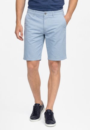 Męski model noszący jasnoniebieskie szorty chino z przednimi kieszeniami, granatowe sneakersy oraz granatową koszulkę, stojący na białym tle.