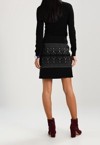 Schwarzer Pullover kombiniert mit einem schwarzen Rock mit einem strukturierten, gemusterten Saum. Das Outfit wird mit burgunderfarbenen Ankle Boots abgerundet.