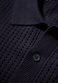 Marinblå pikétröja med ett strukturerat stickmönster, med en svart knapp vid kragen. Tyget är andningsbart med en subtil rutdesign.