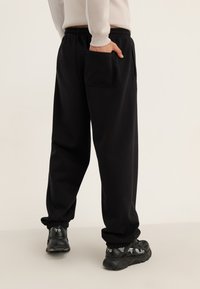 Person iført sorte joggerbukser med elastiske manchetter og sorte sneakers, med hånden i baglommen, stående på et ensfarvet lyst gulv.