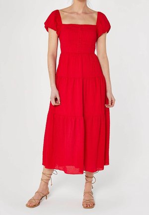Femme portant une robe midi rouge à manches courtes boutonnée, avec un décolleté carré et des sandales à talons beiges à lanières, debout sur un fond blanc.