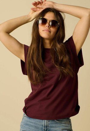 Jeune femme aux longs cheveux bruns portant des lunettes de soleil rondes, un t-shirt bordeaux et un jean bleu clair, posant les bras levés au-dessus de la tête.