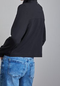 Svart kort jacka med hög krage och slät textur, parat med ljusblå denimjeans med synliga bakfickor.