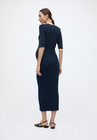 Φόρεμα πλεκτό ρίμπ ναυτικού χρώματος με μανίκια μέχρι τον αγκώνα και κυματιστά τελειώματα, εφαρμοστή γραμμή, μήκος μέχρι τη μέση της γάμπας, συνδυασμένο με μαύρα φλατ παπούτσια.