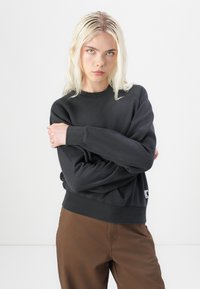 Hanorac negru dintr-un material moale, cu guler rotund și manșete ribbate. Prezintă o etichetă cu logo mic pe tiv. Asortat cu pantaloni maro.