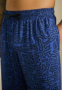 Zilie šorti ar melnu abstraktu rakstu, elastīgu jostasvietu un aukliņas detaļu. Izgatavoti no viegla auduma ar gludu tekstūru.