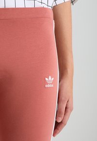 Rosa bomullleggings med en mjuk textur, med en vit adidas-logotyp och sidosträck i vitt. Tajt design utan synlig hårdvara.