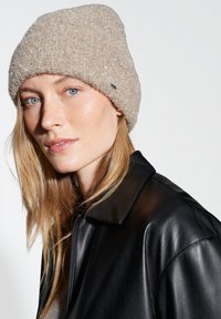 Beige fuzzy knit beanie with a soft texture and small branded tag, worn by a person with shoulder-length hair, against a light background.