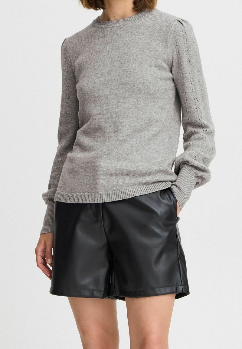 Femme portant un pull en maille gris clair avec des manches à motifs et un short en cuir noir jusqu'aux genoux, debout avec une main dans la poche.
