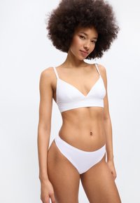 Femme aux cheveux bouclés naturels portant un soutien-gorge blanc et une culotte blanche assortie, debout devant un fond blanc uni.