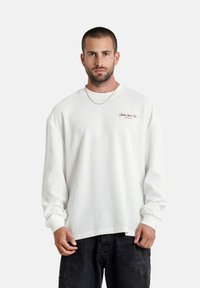 Man met kort haar en een baard draagt een witte shirt met lange mouwen en een textuur, met een klein rood logo op de borst en zwarte broek, staande tegen een witte achtergrond.