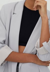 Veste grise claire au design oversize avec manches retroussées, associée à un crop top côtelé noir. Texture lisse et détails minimaux.