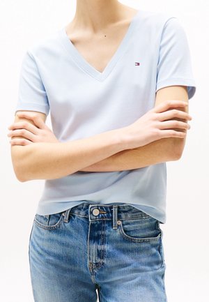 Person wearing light blue V-neck short-sleeve shirt with small logo, crossed arms, and blue denim jeans.