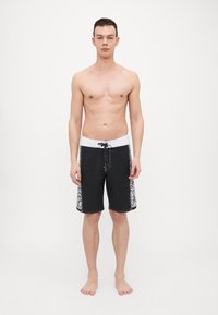 Svart badetights med hvit midje og snøring, med mønstrede sidestykker. Materialet er lett og designet for vannaktiviteter.