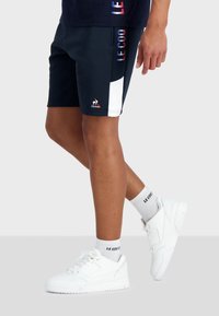Pantalones cortos deportivos en color navy con un panel lateral blanco y logo. Combinados con zapatillas blancas y calcetines estampados. Enfoque en el diseño deportivo y el material.