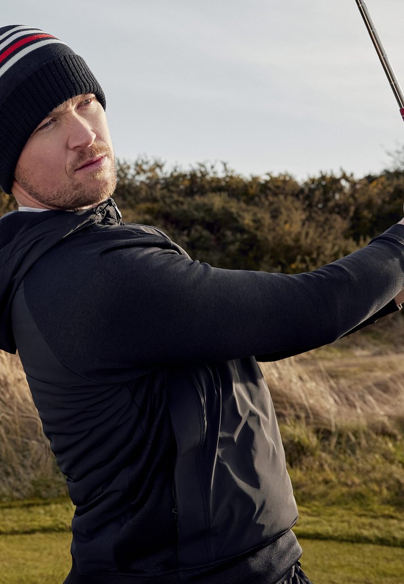 Schwarze Hybrid-Golfjacke mit strukturierten Ärmeln und taillierter Passform, kombiniert mit einer schwarzen Mütze mit roten und weißen Streifen.