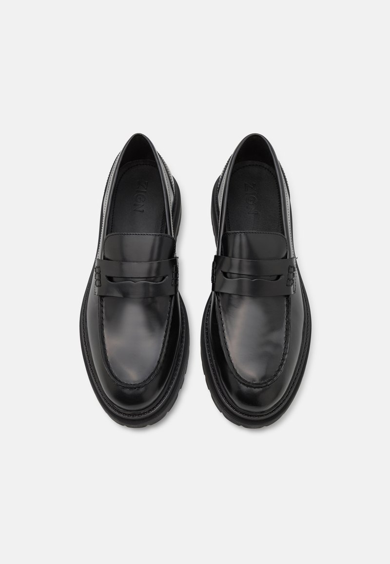 Une paire de mocassins en cuir noir brillant avec des bouts ronds et une sangle décorative, vue de dessus sur un fond blanc.