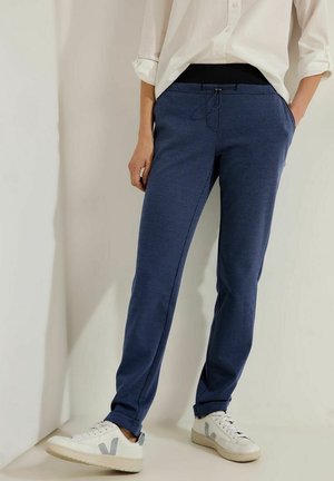 Personne portant un pantalon slim à carreaux bleus avec une ceinture élastique noire, une chemise blanche retroussée et des baskets blanches, debout les mains dans les poches.