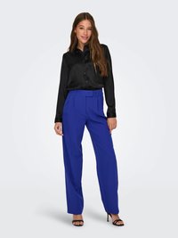 Schwarzes Satin-Hemd mit Knöpfen, kombiniert mit hellblauen hoch taillierten Hosen; das Model trägt schwarze Riemchenschuhe. Glatte Textur und taillierte Passform.