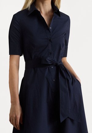 Femme portant une robe boutonnée bleu marine foncé à manches courtes avec un col et une ceinture nouée à la taille.