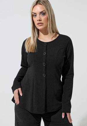 Kobieta o długich blond włosach, ubrana w czarną, zapinaną na guziki bluzkę z długim rękawem i czarne spodnie, pozująca na jasnym tle.