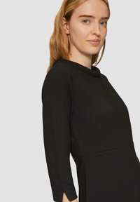 Robe noire avec un motif texturé, col drapé et manches trois-quarts, présentant un corsage ajusté et une jupe évasée.