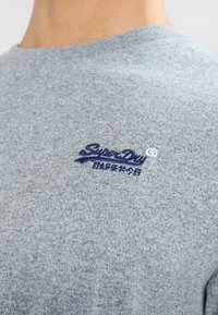 Vaaleanharmaa collegepaita, jossa on rakenteellinen pintakuvio. Sininen brodeerattu logo. Pyöreä kaula-aukko, pehmeä kangas ja lyhyet hihat.