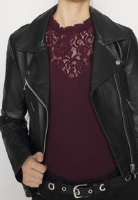 Giacca biker in pelle nera sopra un top bordeaux con dettagli in pizzo e scollatura a scallop. Cintura nera con inserti in metallo argentato.