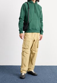 Vihreä huppari kenguruntaskulla, yhdistetty beigen värisiin monitaskullisiin cargo-housuihin ja mustiin lenkkareihin, joissa on valkoiset nauhat.