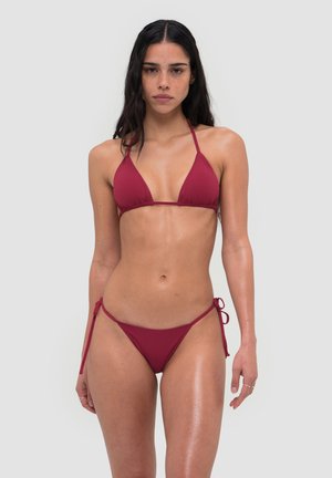 Femme aux longs cheveux foncés portant un bikini rouge noué, debout devant un fond uni gris clair, face à la caméra avec une expression neutre.