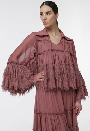 Mujer de cabello oscuro recogido en una coleta baja lleva un vestido lila de encaje y tul de capas con mangas anchas de volantes, de pie ante un fondo liso.