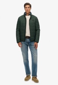 Grøn quiltet jakke med høj krave, hvid tekstureret sweater, lyseblå jeans og beige sko. Enkel design, ingen synlige mønstre eller logoer.