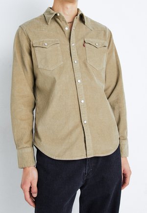 Homme portant une chemise beige en velours côtelé à boutons-pression argentés avec poches à rabat, associée à un jean noir et un collier à chaîne.